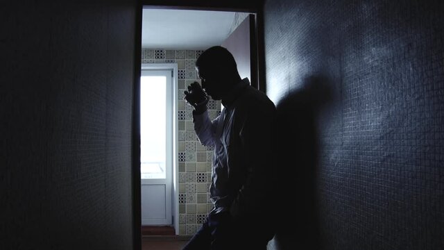A man drinking coffee and talking aloud to himself wearing a white t-shirt leaning his back against the wall in the hallway of a soviet apartment , depressed mood, cramped dark room, lonely person.