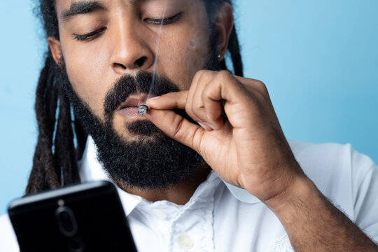 Black  man with dreadlocks and black beard smoking weed holding the cell blue bottom,  banground ,Marijuana joint in the hand, Concepts of medical marijuana use and legalization of the cannabis. tecno