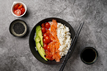 Poke bowl - black bowl with white rice, smoked salmon, cherry tomatoes and avocado, sesame seeds, soy sauce and chopsticks on a grey concrete surface, top view