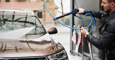 Cropped view of young man washing car on carwash station outdoor. Handsome worker cleaning automobile, using high pressure water.