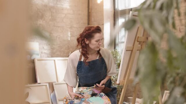 Tracking of young red-haired Caucasian woman wearing apron, entering art studio, sitting down and painting on canvas at daytime