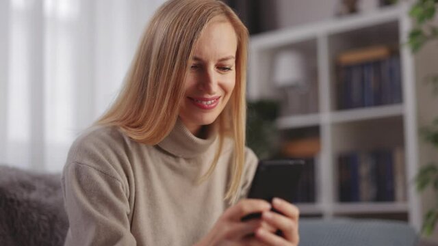 Positive caucasian woman typing messages on modern smartphone while resting on comfy couch. Online communication during free time at modern apartment.