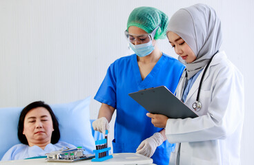 Asian female nurse in face mask safety goggles blue uniform collect blood sample while Muslim Islam Arab doctor in white lab coat with hijab stethoscope stand smile while patient sleep