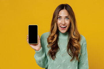 Young smiling amazed woman 30s in green knitted sweater hold use mobile cell phone with blank screen workspace area isolated on plain yellow color background studio portrait. People lifestyle concept.