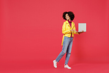Full size body length bright happy fancy young curly black latin woman 20s wears yellow jacket go move hold use work on laptop pc computer look back isolated on plain red background studio portrait.