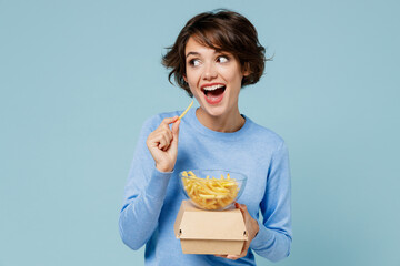 Young smiling cheerful fun cool woman 20s in sweater hold burger french fries potato look aside on...