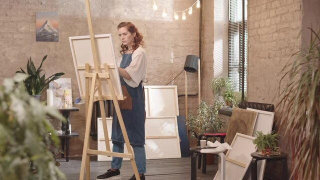 Full shot of young red-haired Caucasian woman wearing apron, standing and painting on canvas at daytime in art studio