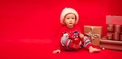 Little funny Santa with a Christmas tree toy in his hands sits next to Christmas presents on a red background, copy space