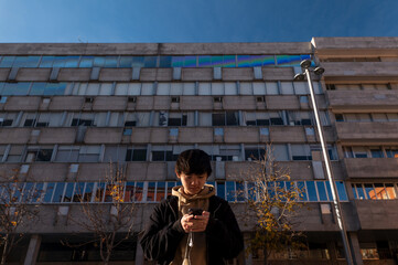 Asian teenage boy looking at mobile phone against building on street. Madrid. Spain