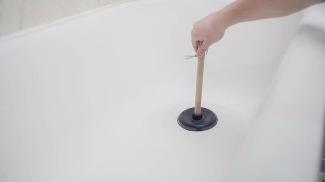 Trying to remove a blockage in a bathroom downpipe with a plunger. A hand uses a plunger in a bathtub with non-flowing water
