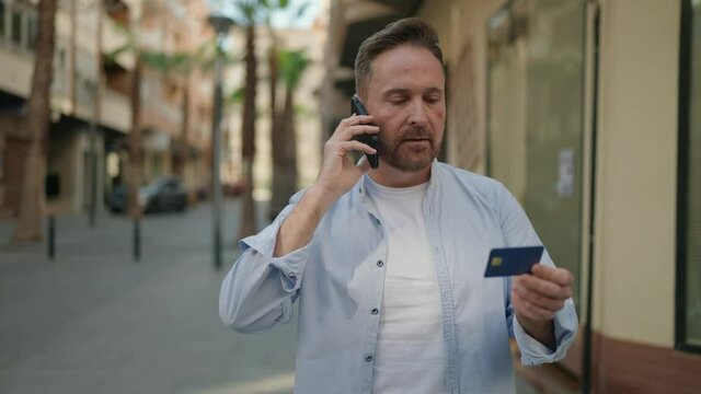 Young caucasian man talking on the smartphone holding credit card at street
