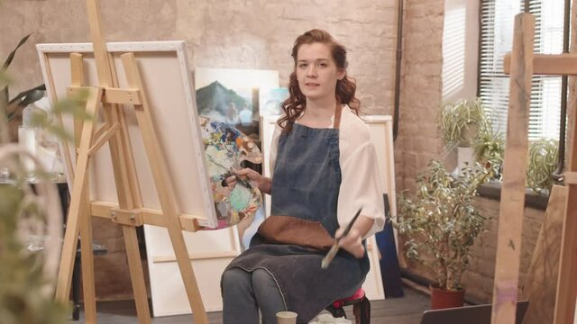 Medium long POV of young Caucasian woman wearing apron, sitting and painting in studio, talking, smiling and showing her artwork on camera