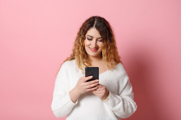Fototapeta premium A cheerful young woman, standing isolated on a pink background with a smartphone in her hands, looks into the camera and smiles.