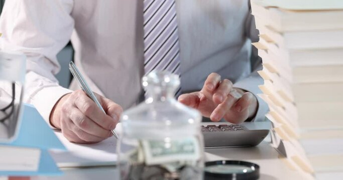 Businessman counting on calculator and writing on paper in front of glass jar with money closeup 4k movie slow motio