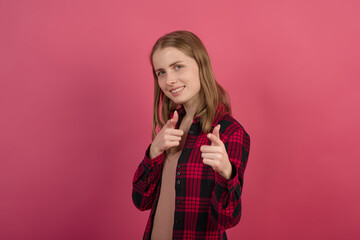 A young and attractive Caucasian blonde girl in a shirt pointing her fingers with pistols at the camera on a pink studio background.