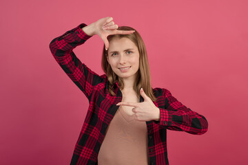 A young and attractive Caucasian blonde girl in a shirt making a camera frame with her hands as if taking pictures on a pink studio background.