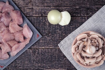Meat cut into pieces, peeled onion and a bowl of chopped champignons on an old wood texture background. Flat lay