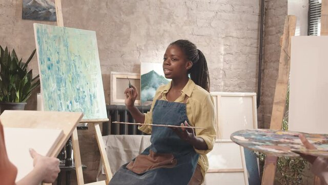 Tracking out of young African American female art teacher wearing apron, showing abstract painting and talking to unrecognizable students in studio
