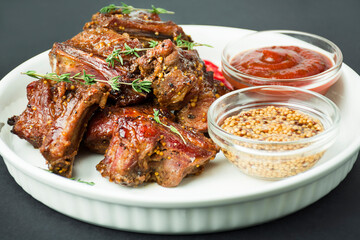 Baked ribs on a white dish. Grilled pork ribs with spices and herbs on a dark background. Food background. Side view.
