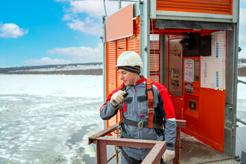 An engineer worker in overalls at a construction site looks down from a high-rise construction site against the background of a construction elevator, talks on a walkie-talkie.