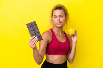 Girl with curly hair isolated on yellow background having doubts while taking a chocolate tablet in one hand and an apple in the other