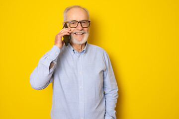 Portrait of happy european senior man talking on phone and smiling isolated over yellow background.