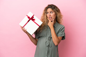 Girl with curly hair isolated on pink background pregnant and holding a gift while having doubts