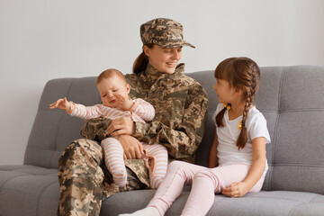 Portrait of smiling soldier woman wearing camouflage uniform sitting on sofa with kids, being...