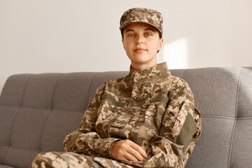 Indoor shot of serious young adult soldier woman wearing camouflage uniform sitting on cough and...