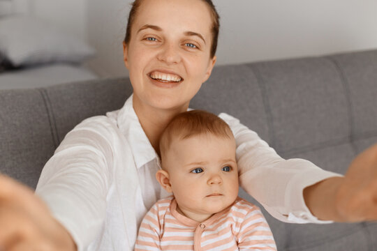 Smiling woman making selfie with her charming infant daughter POV, looking at camera with happy facial expression, point of view photo, female wearing white shirt.