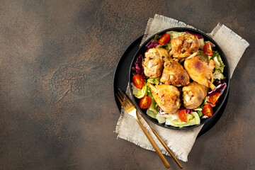Fried chicken legs with salad in a plate on a dark kitchen table. Cooked poultry meat, pickles, and tomatoes on a culinary background top view