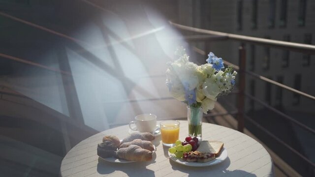 Perfect morning breakfast on balcony with tasty croissants, fresh juice, coffee and fruits. Sunny warm day. Sunlight in footage.