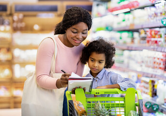 Millennial black mother and her adorable daughter checking grocery list in notebook while shopping...