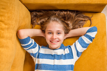A close-up portrait of a smiling beautiful little blonde girl lying on a yellow sofa, a photo on...
