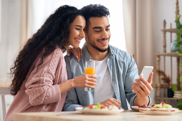 Happy Middle Eastern Couple Browsing Internet On Smartphone While Having Breakfast