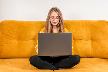 A cute little girl in a sweater uses a laptop for online education and doing school homework or video calls to friends while sitting on a yellow sofa