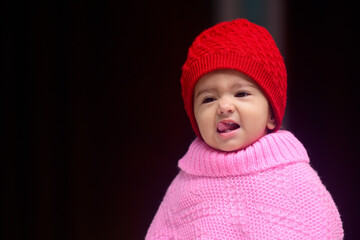 13 months old Indian baby dressed in red winter cap and pink pohcho giving cute expressions while taking her tounge out.
