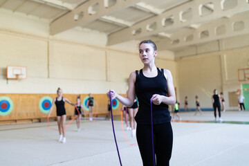Girl gymnast jumping with rope on training in gym with other trainees