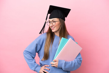 Young student woman wearing a graduate hat isolated on pink background posing with arms at hip and smiling