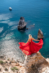 A girl with loose hair in a long red dress descends the stairs between the yellow rocks overlooking the sea. A rock can be seen in the sea. Sunny path on the sea from the rising sun