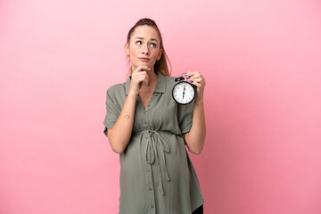 Fototapeta premium Young woman isolated on pink background pregnant and holding clock while thinking