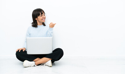 Young mixed race woman sitting on the floor with laptop isolated on white background pointing to the side to present a product