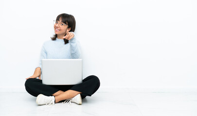 Young mixed race woman sitting on the floor with laptop isolated on white background points finger at you with a confident expression