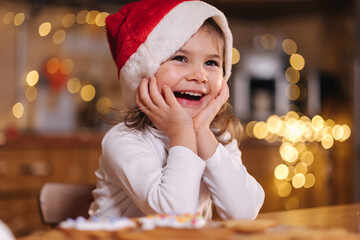 Portrait of beautiful tree year girl in santa hat sitting by the table in front of kitchen...