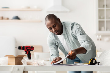 Concentrated african american man reading furniture installation instruction, assembling kitchen...