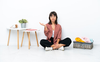 Young mixed race woman folding clothes sitting on the floor isolated on white background having doubts while raising hands