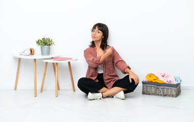 Young mixed race woman folding clothes sitting on the floor isolated on white background pointing back