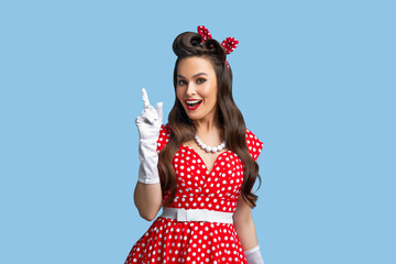 Beautiful young pinup lady in retro style polka dot dress pointing upwards on blue studio background