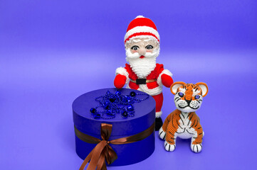 On a purple background, knitted toys of Santa Claus and tiger are standing near a purple gift box.