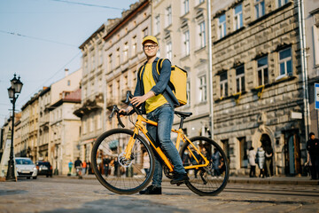 Fototapeta na wymiar Portrait of a happy delivery man who is sitting on a bicycle and looking at the camera. He is carrying a yellow backpack on his shoulders.